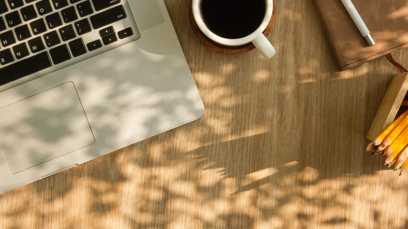 Draufsicht auf Laptop, Kaffeetasse und Tagebuch auf einem Holztisch – mit Platz für Text.